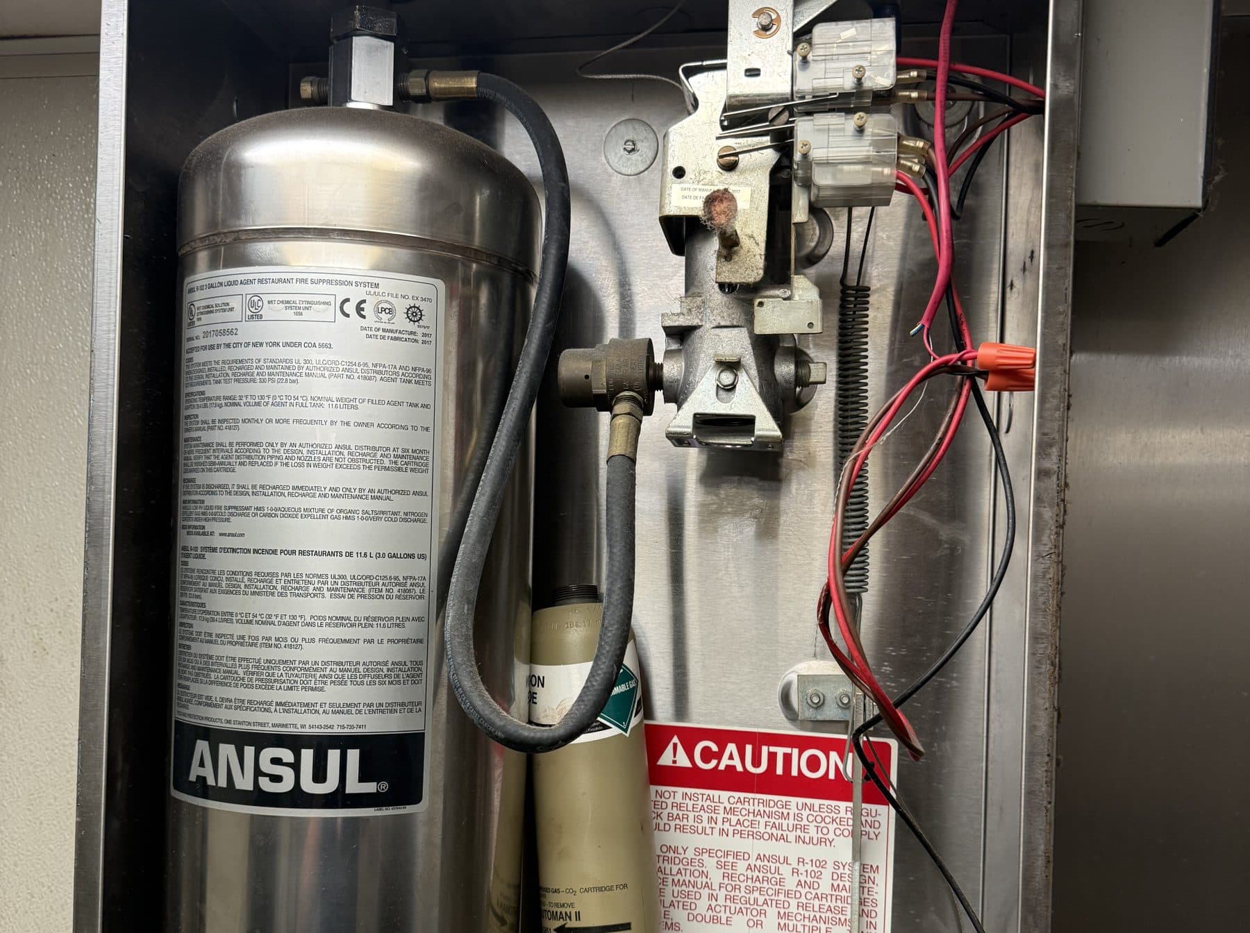 Open ANSUL R-102 release cabinet showing the agent tank, the expellant gas cartridge, the AUTOMAN release and its caution label