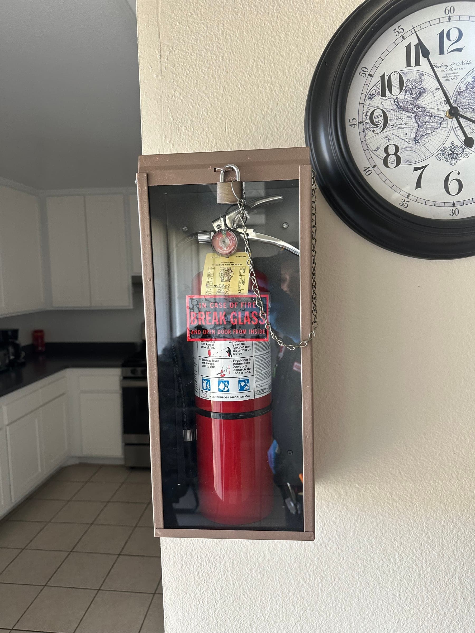 ABC extinguisher in a locked break-glass cabinet on a wall beside a clock, with a fresh State Fire Marshal tag