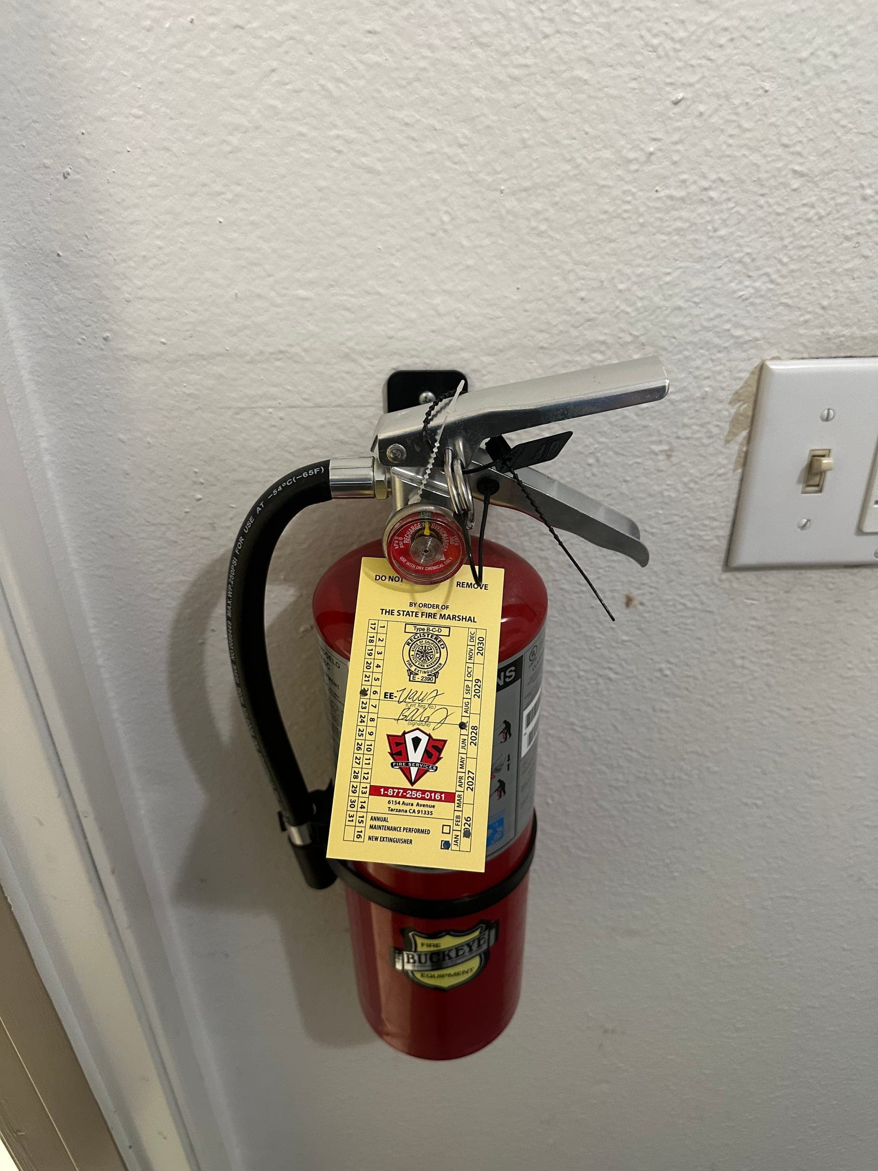 Red 5 lb ABC extinguisher on a heavy-duty wall bracket beside a light switch, tagged for 2026