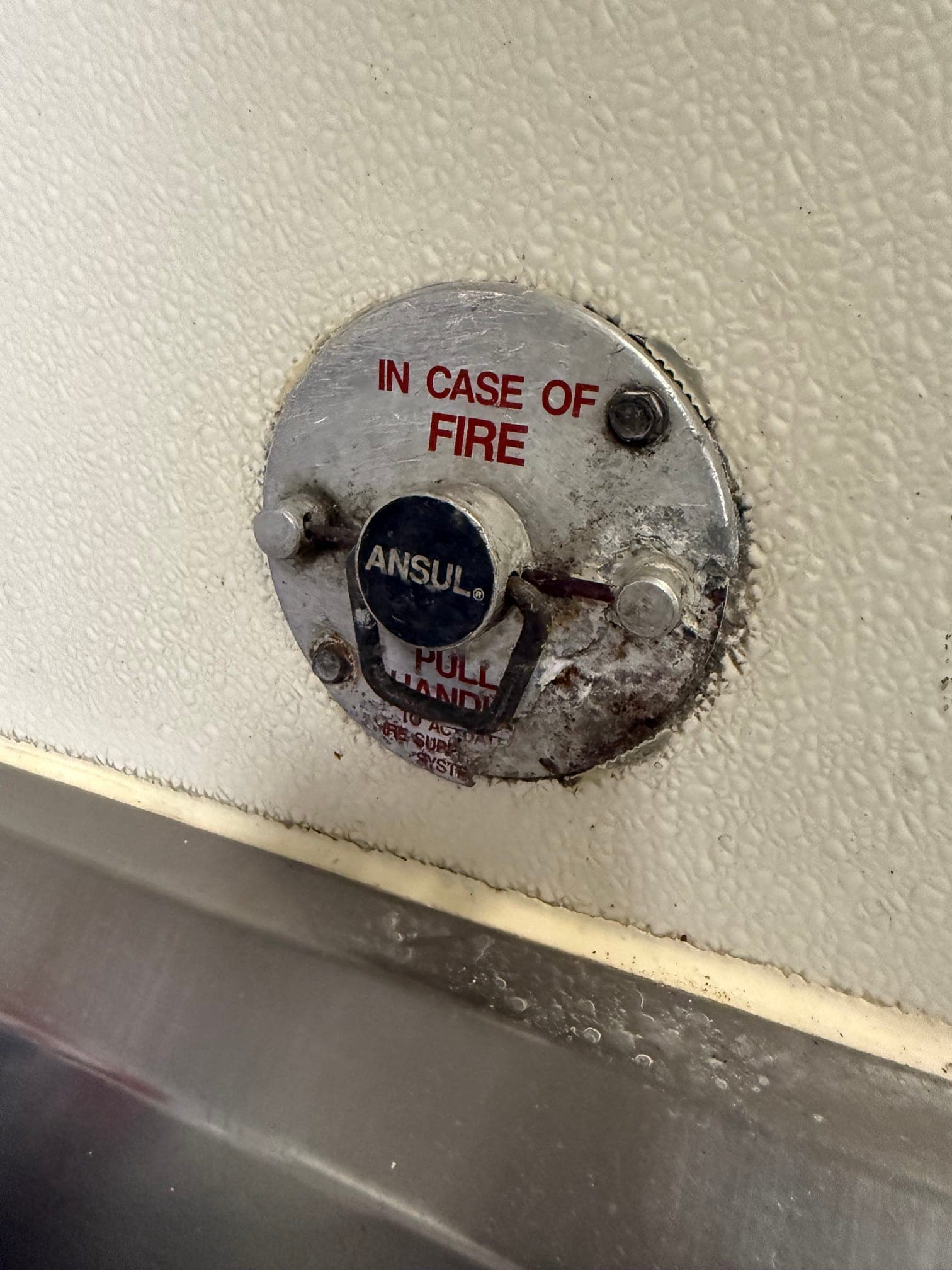 Corroded Ansul manual pull station on a kitchen wall above a stainless sink, its faceplate stained with rust