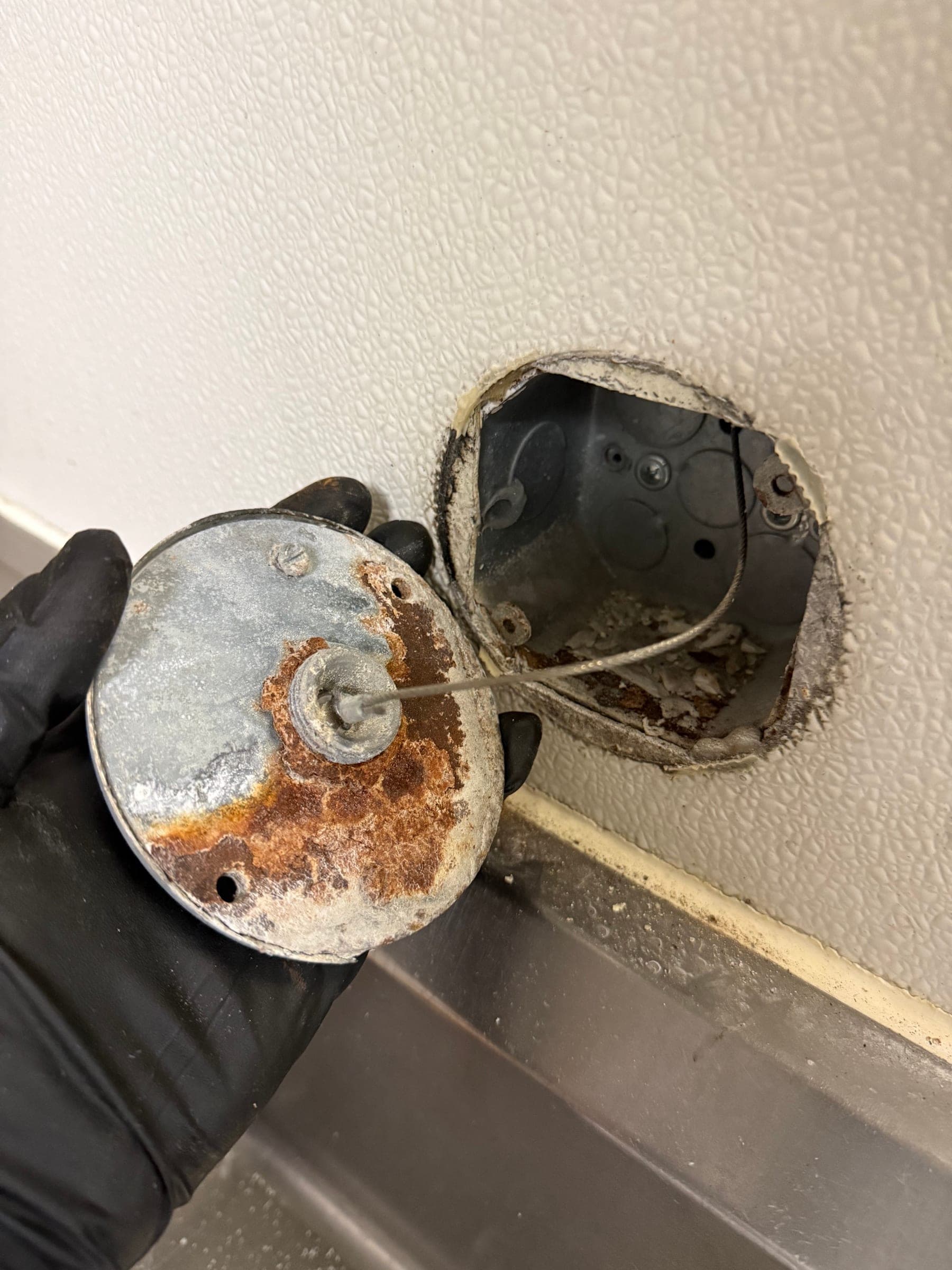 Old faceplate removed and held in a gloved hand, its back rusted through, with the cable running into the wall box
