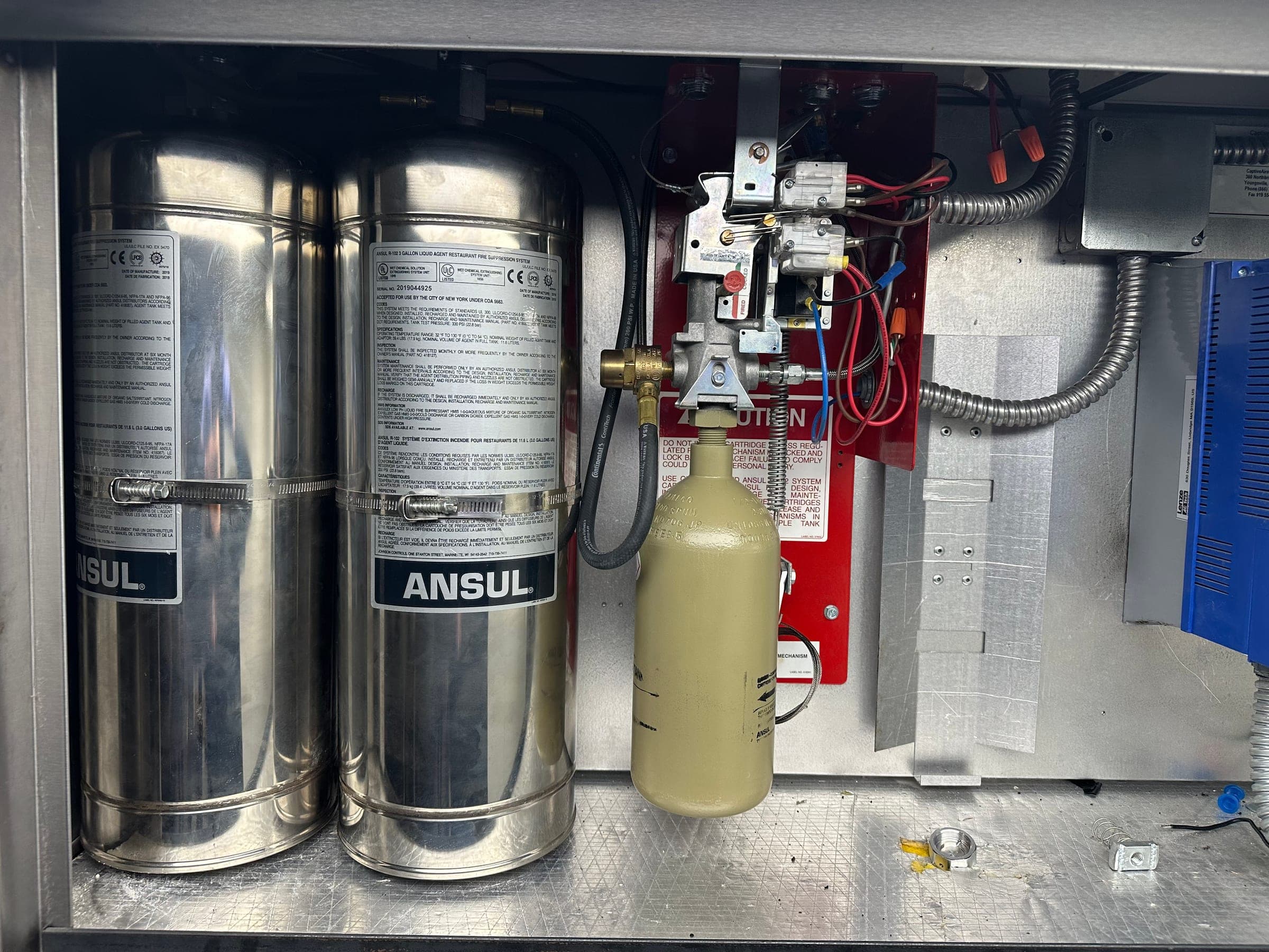 Two stainless Ansul R-102 agent cylinders beside the AUTOMAN release and its expellant cartridge in the hood cabinet