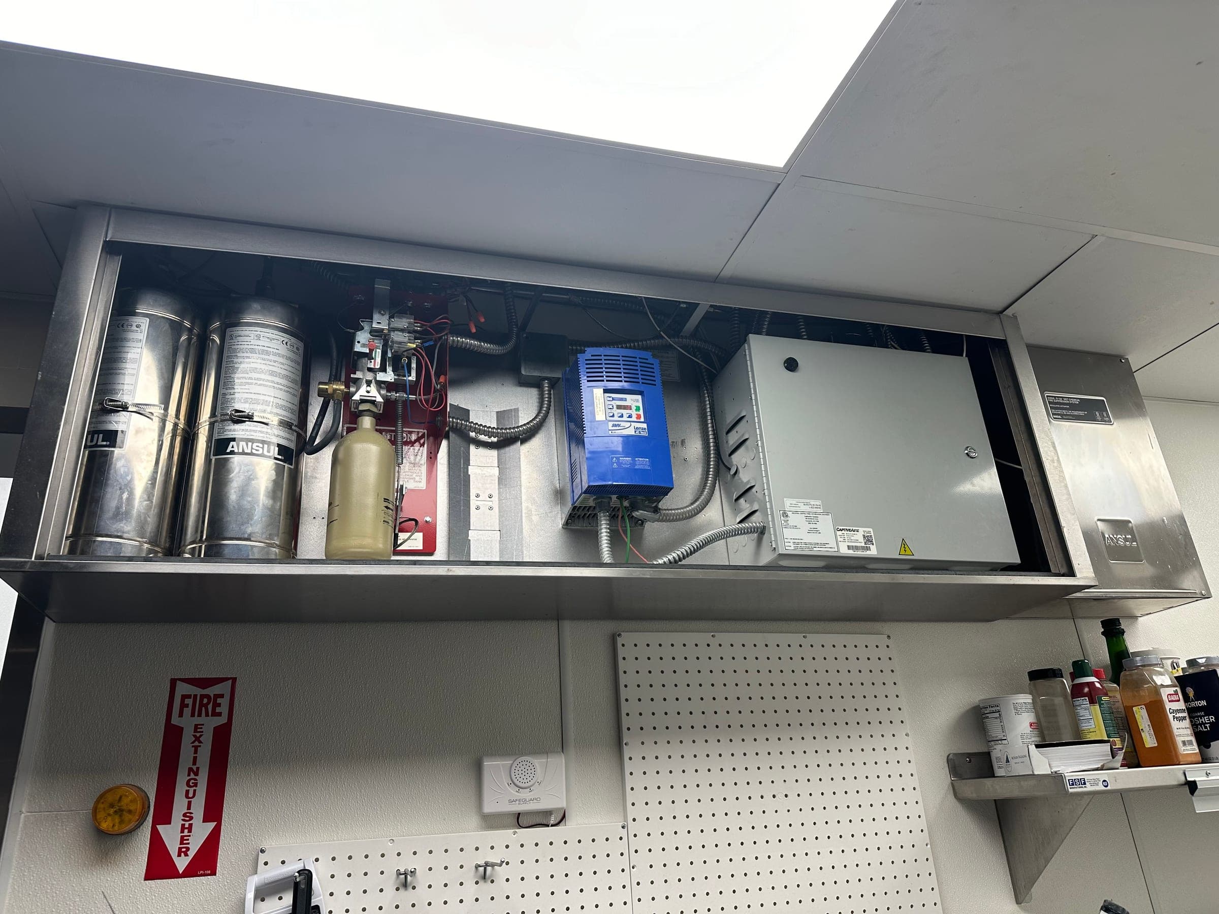 Open utility cabinet above a kitchen line holding Ansul cylinders, the release mechanism, a fan drive and a control box