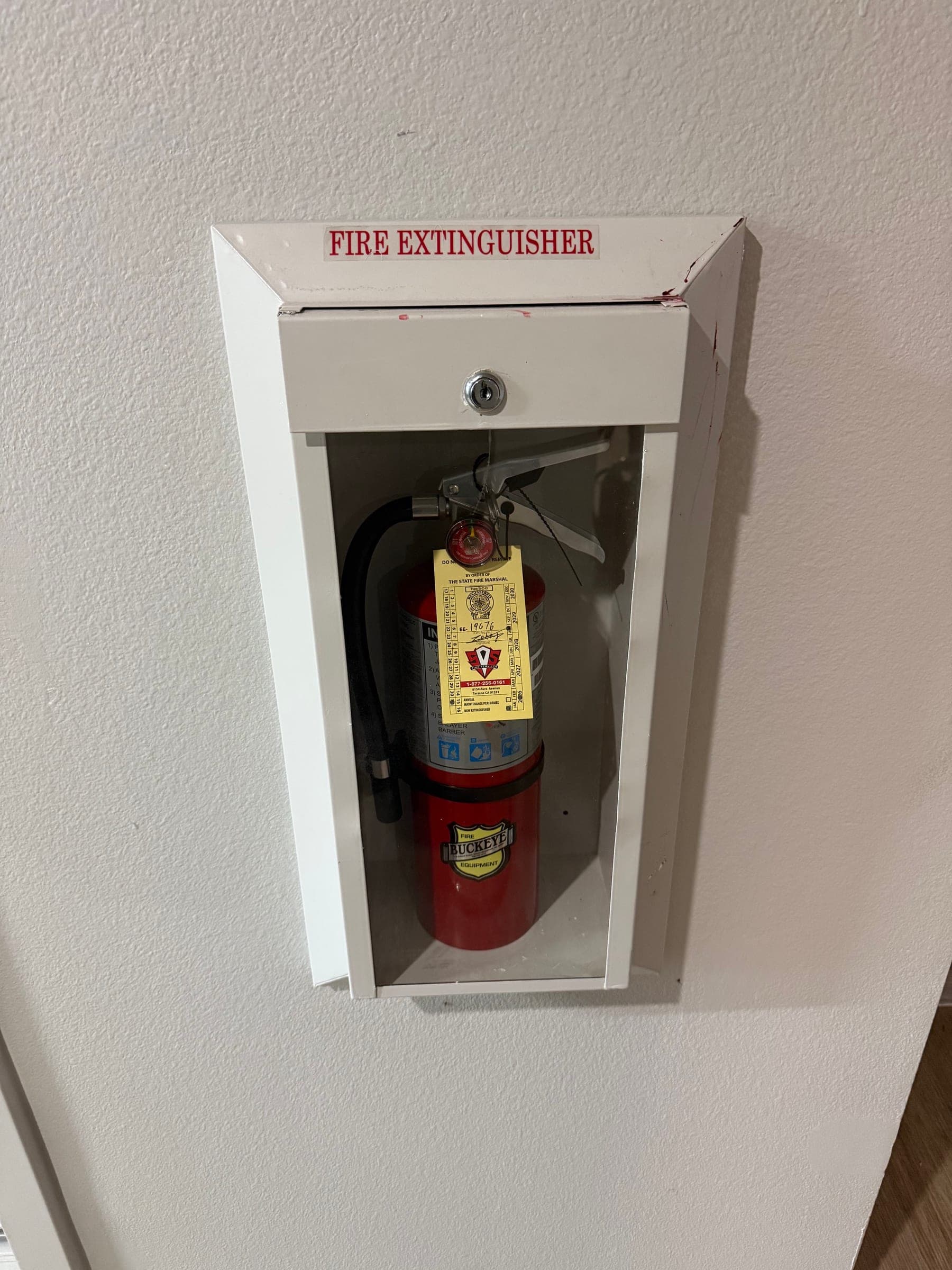 White extinguisher cabinet with a red label and a service-tagged ABC extinguisher behind clear glazing