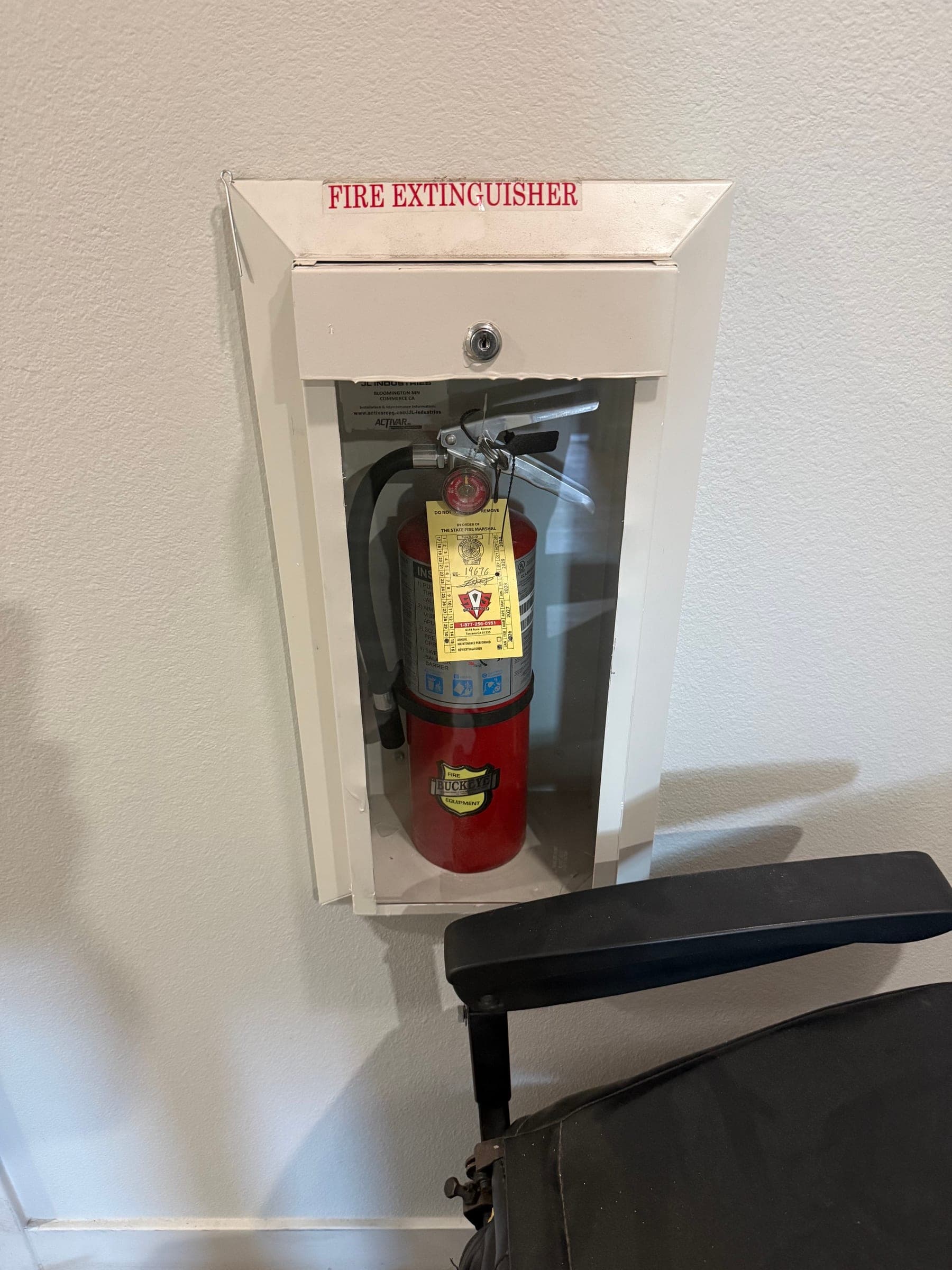 Labeled extinguisher cabinet with a tagged ABC extinguisher, mounted above a chair in a common area