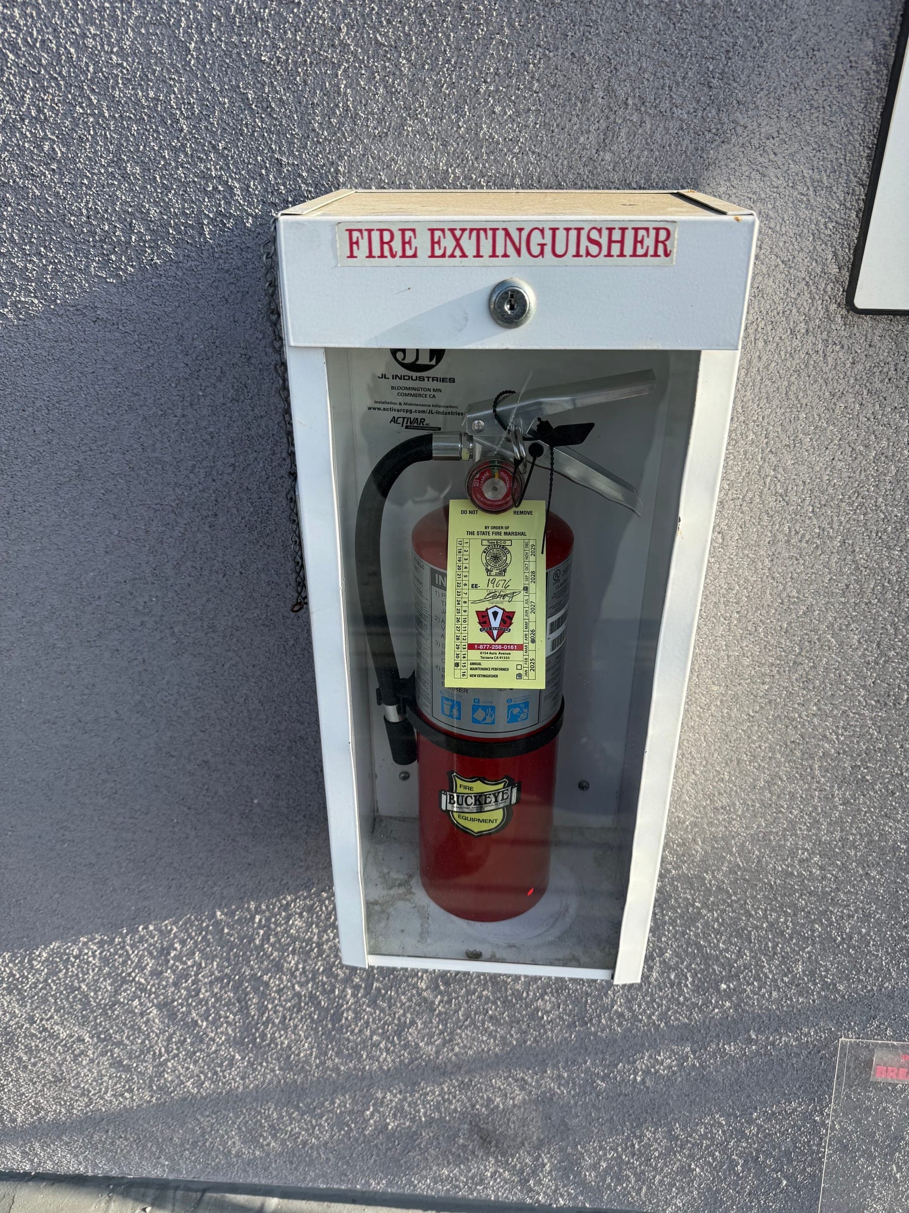 Surface-mounted extinguisher cabinet on a stucco wall with a tagged ABC extinguisher inside