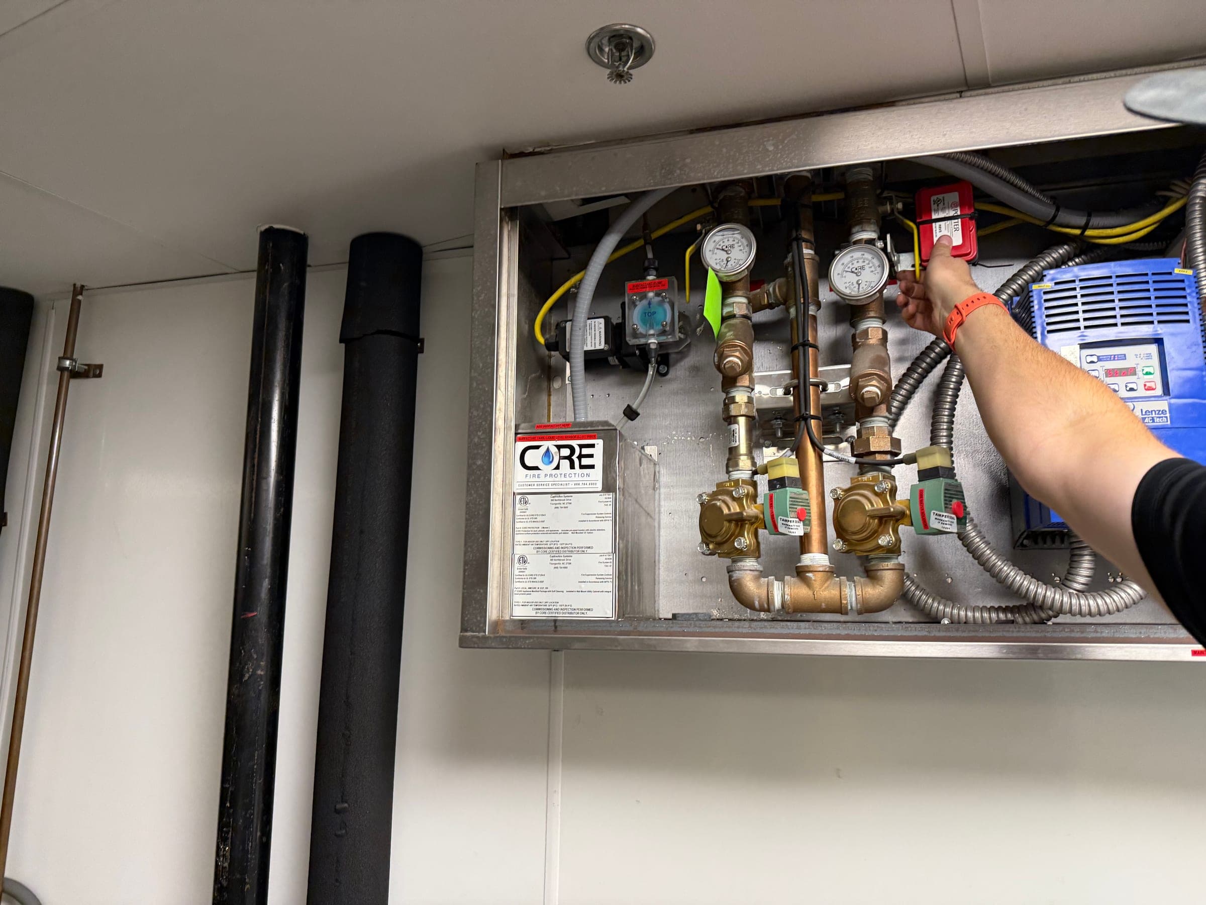 Technician's hand at an open CaptiveAire CORE control cabinet with gauges, solenoid valves and piping