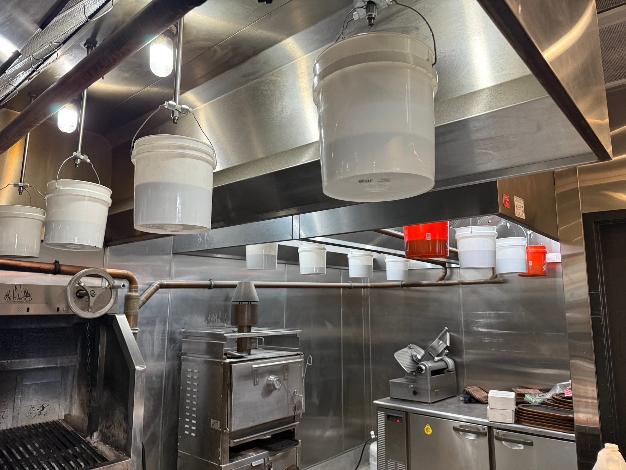 Test buckets hung under hood nozzles over a wood-fired grill line during a semi-annual suppression inspection