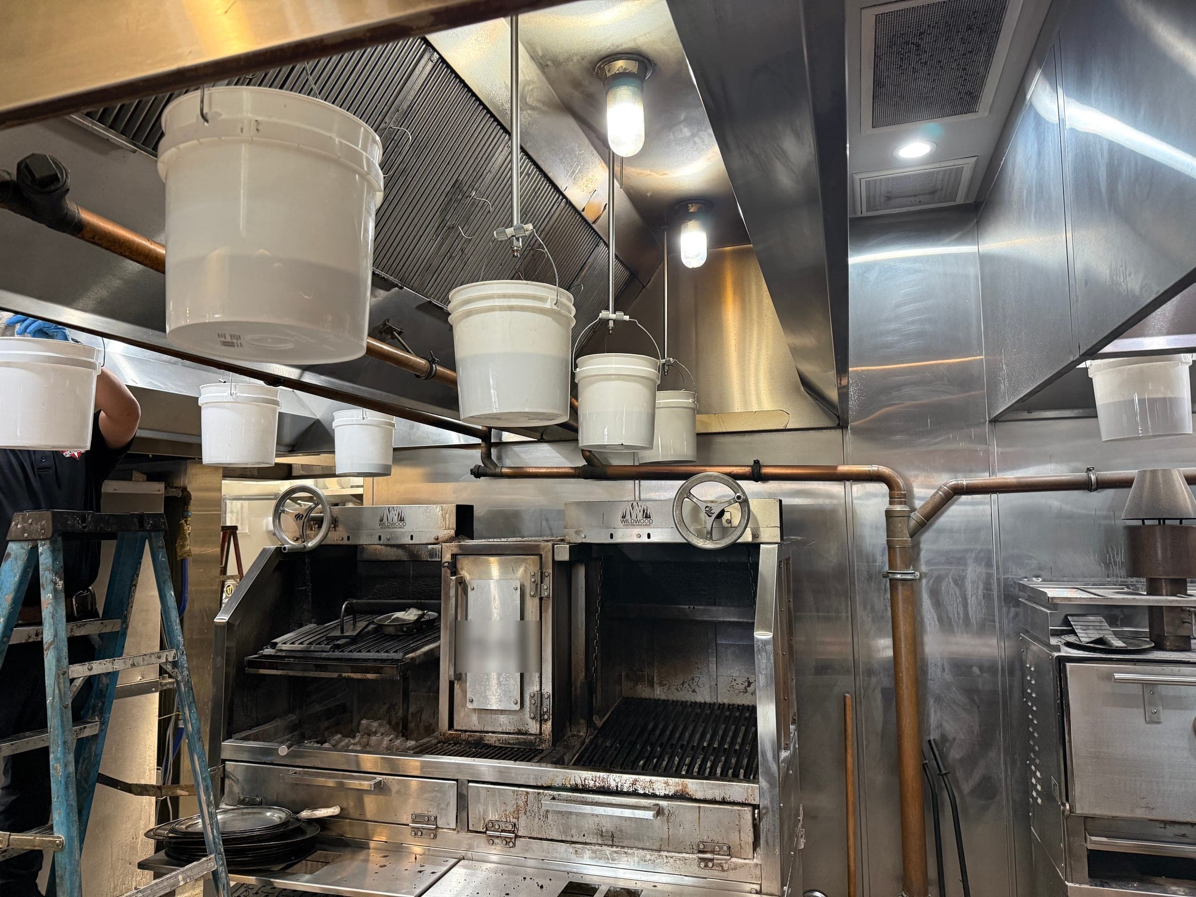 Technician on a ladder setting test buckets under nozzles above a wood-fired grill