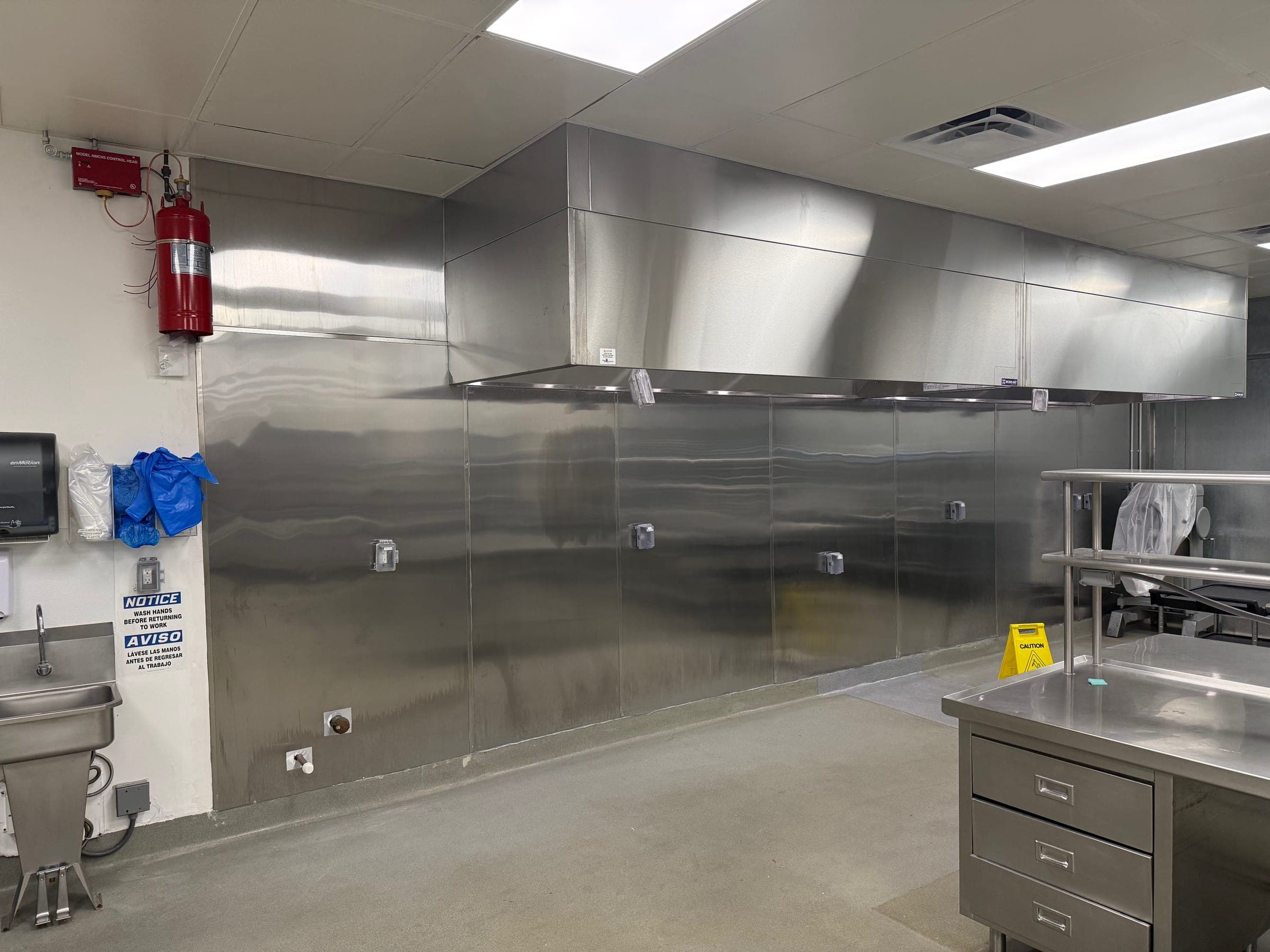Industrial kitchen wall with the Pyro-Chem system mounted beside a long stainless exhaust canopy and stainless wall panels
