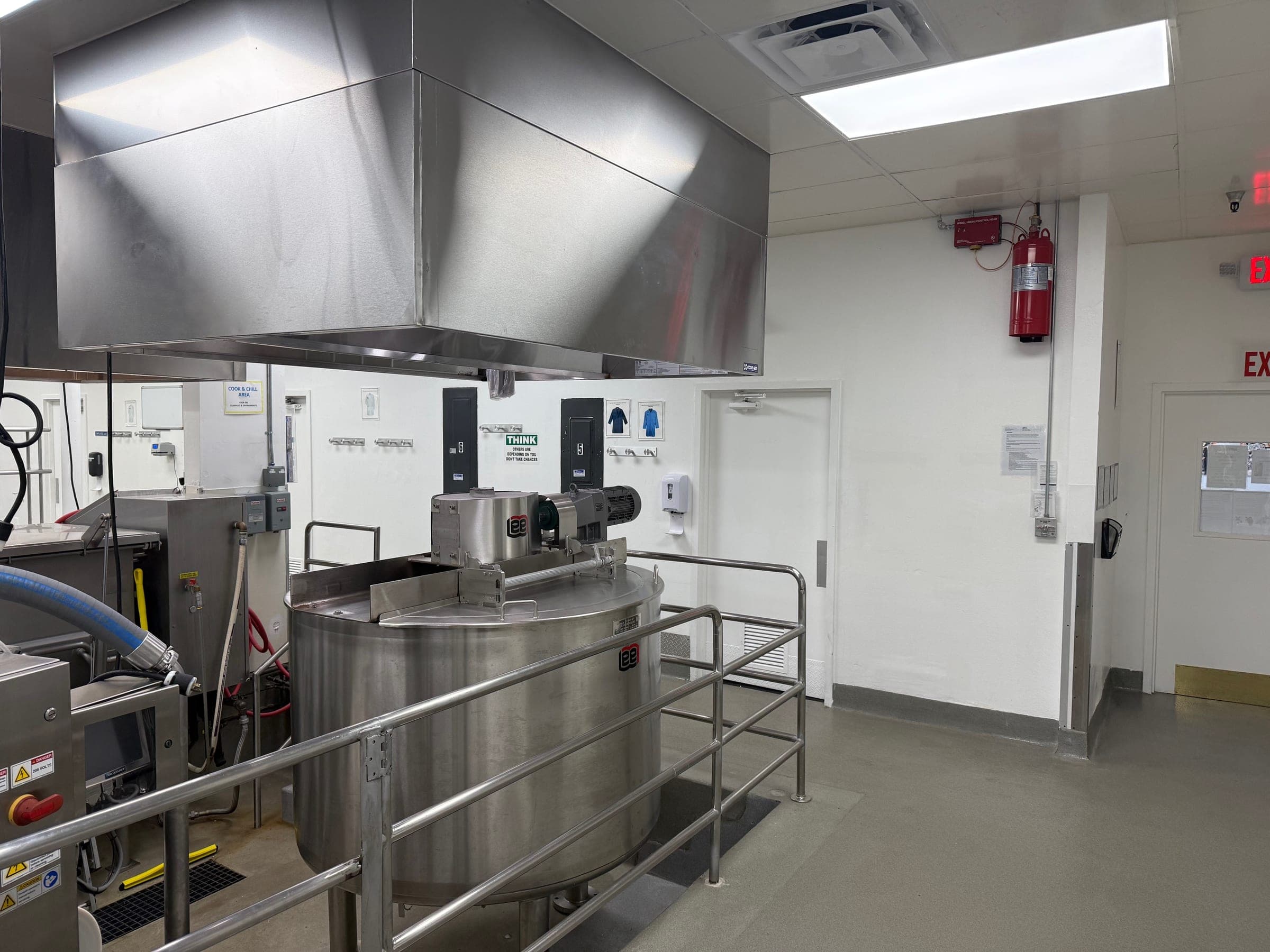 Stainless exhaust canopy over a large cook-and-chill kettle in an industrial kitchen, the suppression cylinder on the far wall
