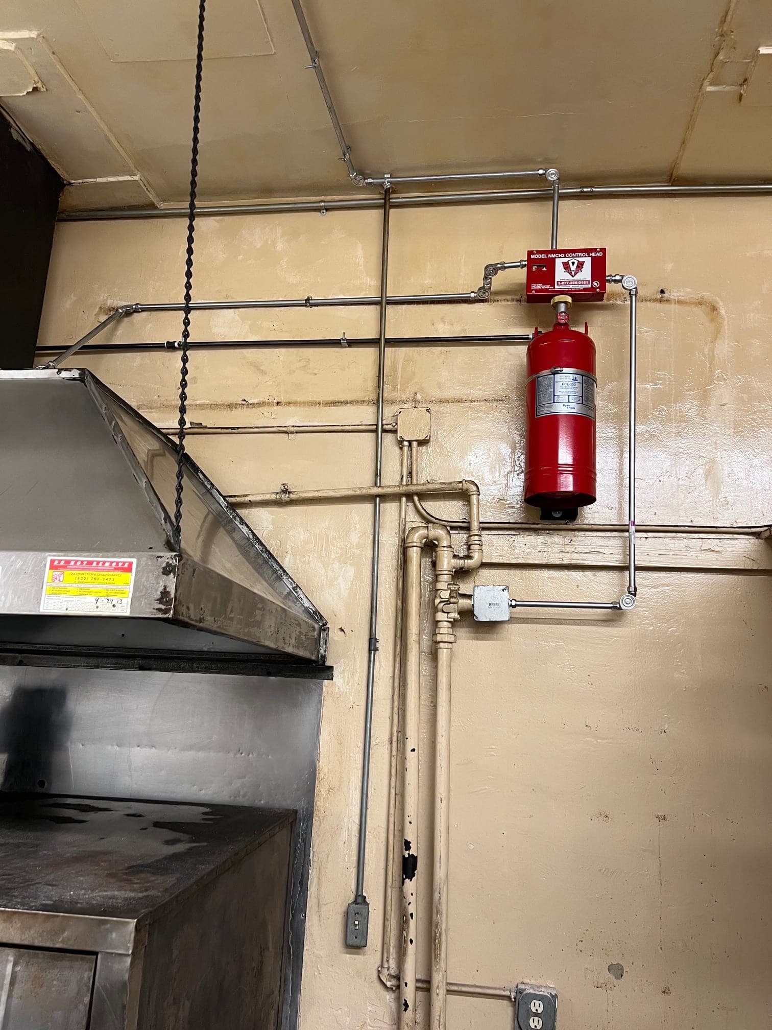 Wall-mounted Pyro-Chem system beside the exhaust hood, with piping running up and across to the hood