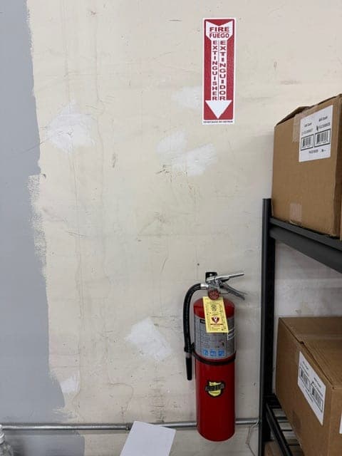 Tagged ABC extinguisher mounted on a patched warehouse wall next to storage shelving, sign above